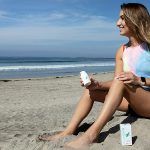 a woman sitting at the beach holding a bottle of Resilience CBD lotion